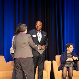 This is a photo of Dr. Kenneth Alleyne speaking to Victoria Veltri on stage at an event while other people are seated behind them.