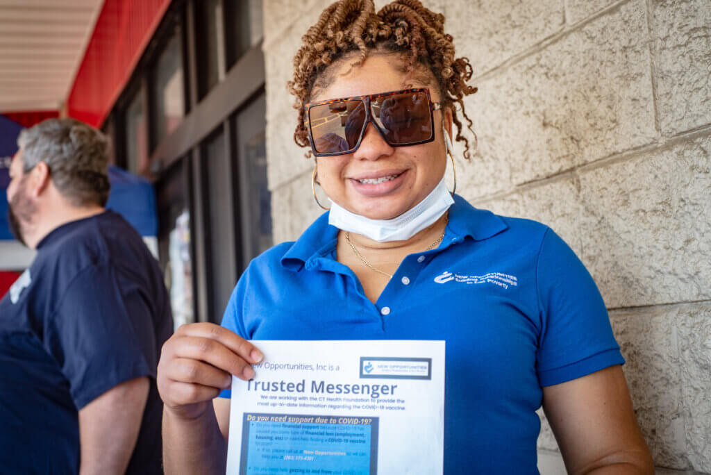 This is a picture of a woman wearing sunglasses and a face mask standing outside holding a paper that says "Trusted messenger."